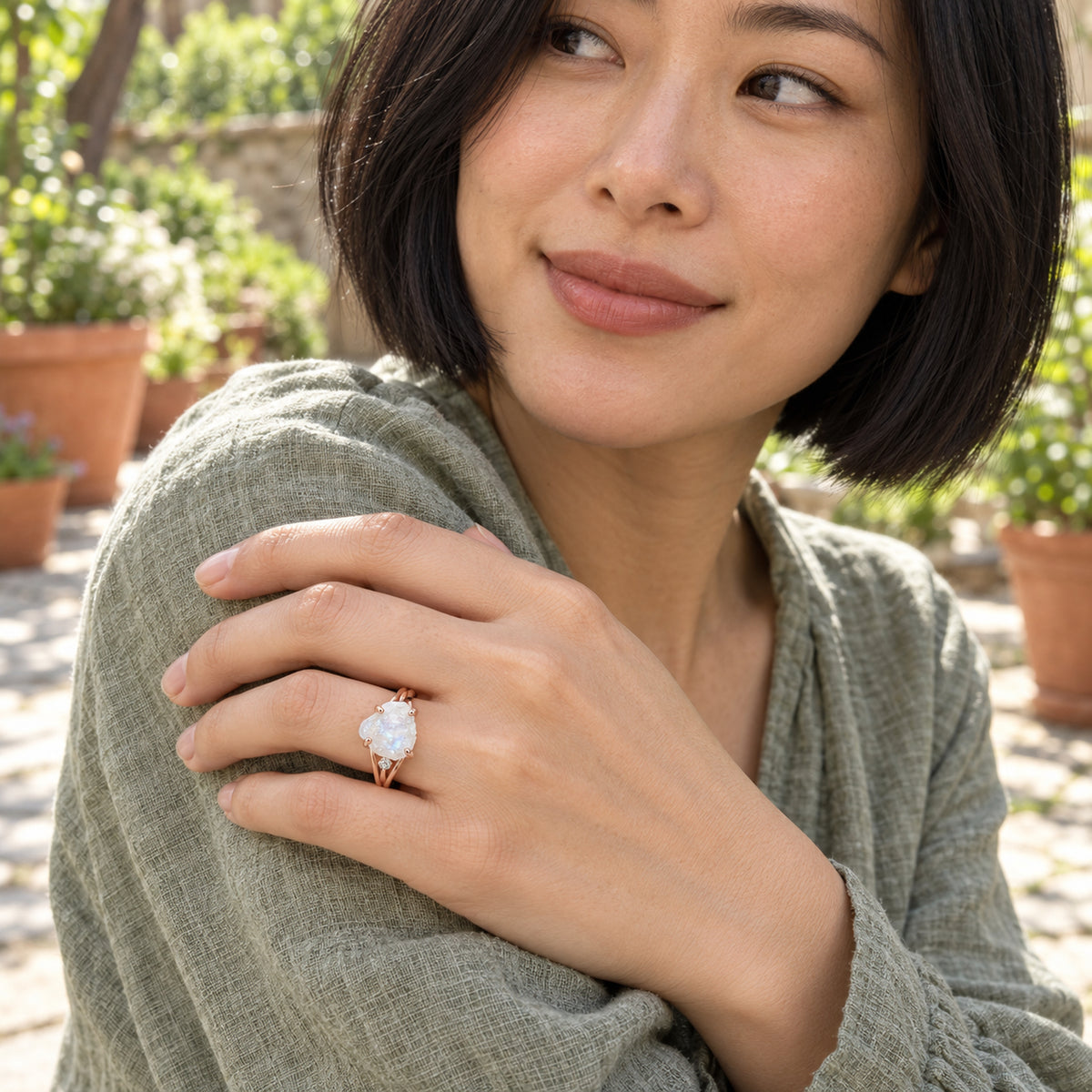 The rose gold moonstone ring is worn on a hand resting against a sage linen sleeve in a leafy courtyard.