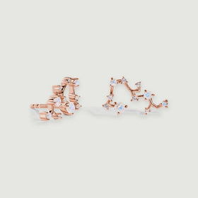Two rose gold constellation stud earrings with round iridescent stones and clear accents rest on a light gray surface.