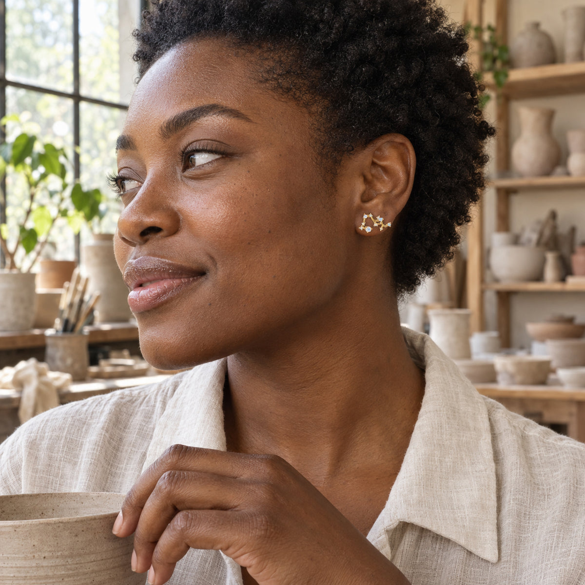 A woman with short natural curls wears a yellow gold constellation stud beside pottery shelves and a workshop window.
