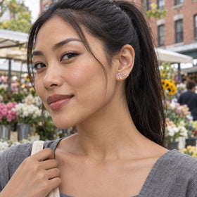 A woman with a black ponytail wears a silver constellation stud with colorful flower stalls and brick buildings behind her.