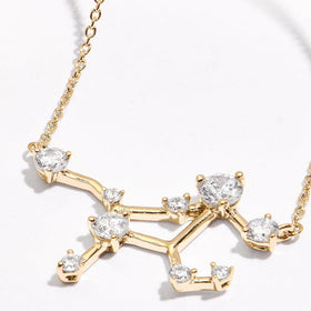 A yellow gold constellation pendant and portions of its chain lie on a pale tabletop in angled light.