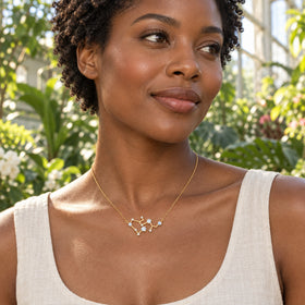 A woman with a dark bob wears a yellow gold branching necklace over a white linen shirt while holding a cup at an outdoor cafe.