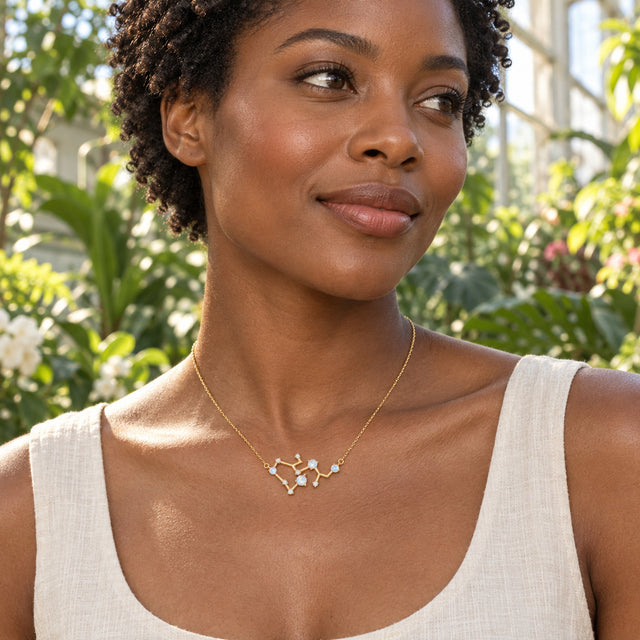 A woman with a dark bob wears a yellow gold branching necklace over a white linen shirt while holding a cup at an outdoor cafe.