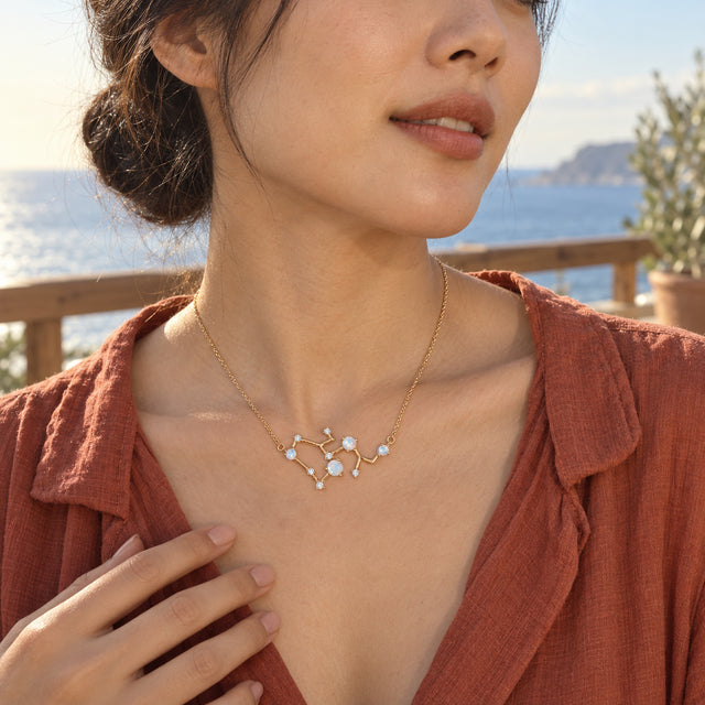 A woman with short natural curls wears a yellow gold necklace with pale round stones and clear accents above a terracotta top in a garden courtyard.