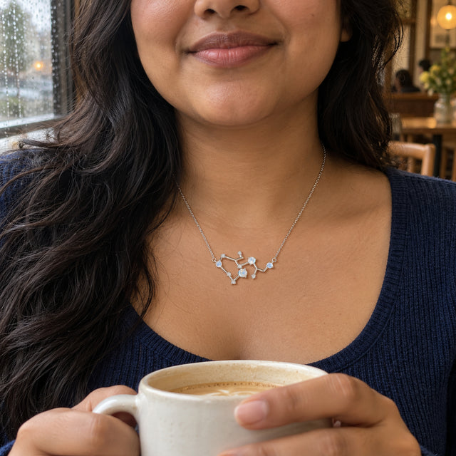 A woman with long dark waves wears a silver constellation necklace with pale blue stones above a navy shirt beside a window and bookshelves.