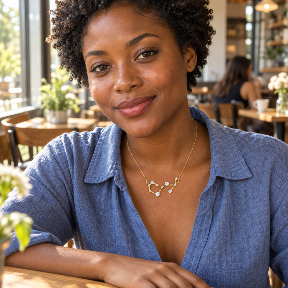A woman in a white linen shirt wears a yellow gold constellation necklace while holding a ceramic cup in a leafy courtyard.