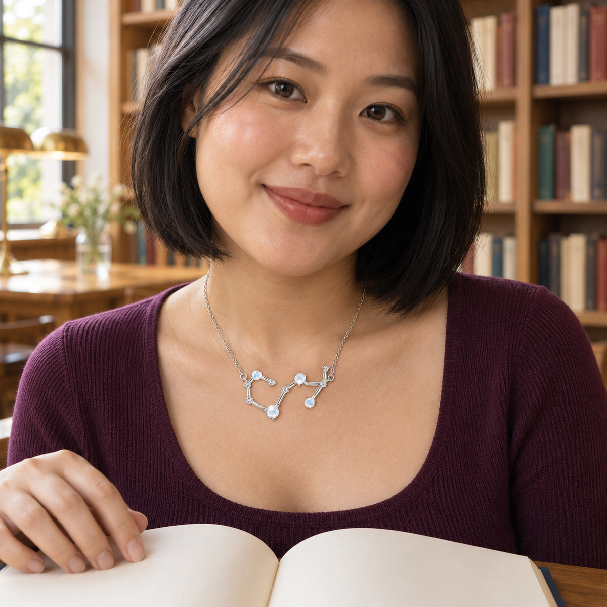 A silver necklace with pale round stones rests below the collarbones of a woman in a blue blouse holding a book beside wooden shelves.