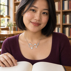 A silver necklace with pale round stones rests below the collarbones of a woman in a blue blouse holding a book beside wooden shelves.
