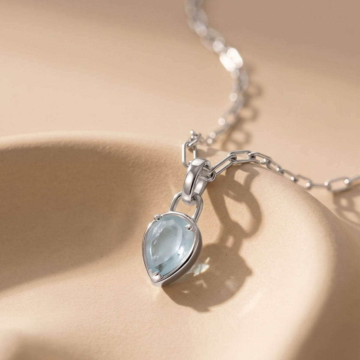 A silver-colored aquamarine pendant rests at an angle on a curved beige ceramic surface with its linked chain behind it.