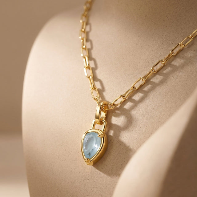 A yellow gold-colored necklace drapes over a beige ceramic curve, showing the pendant edge and elongated chain links.
