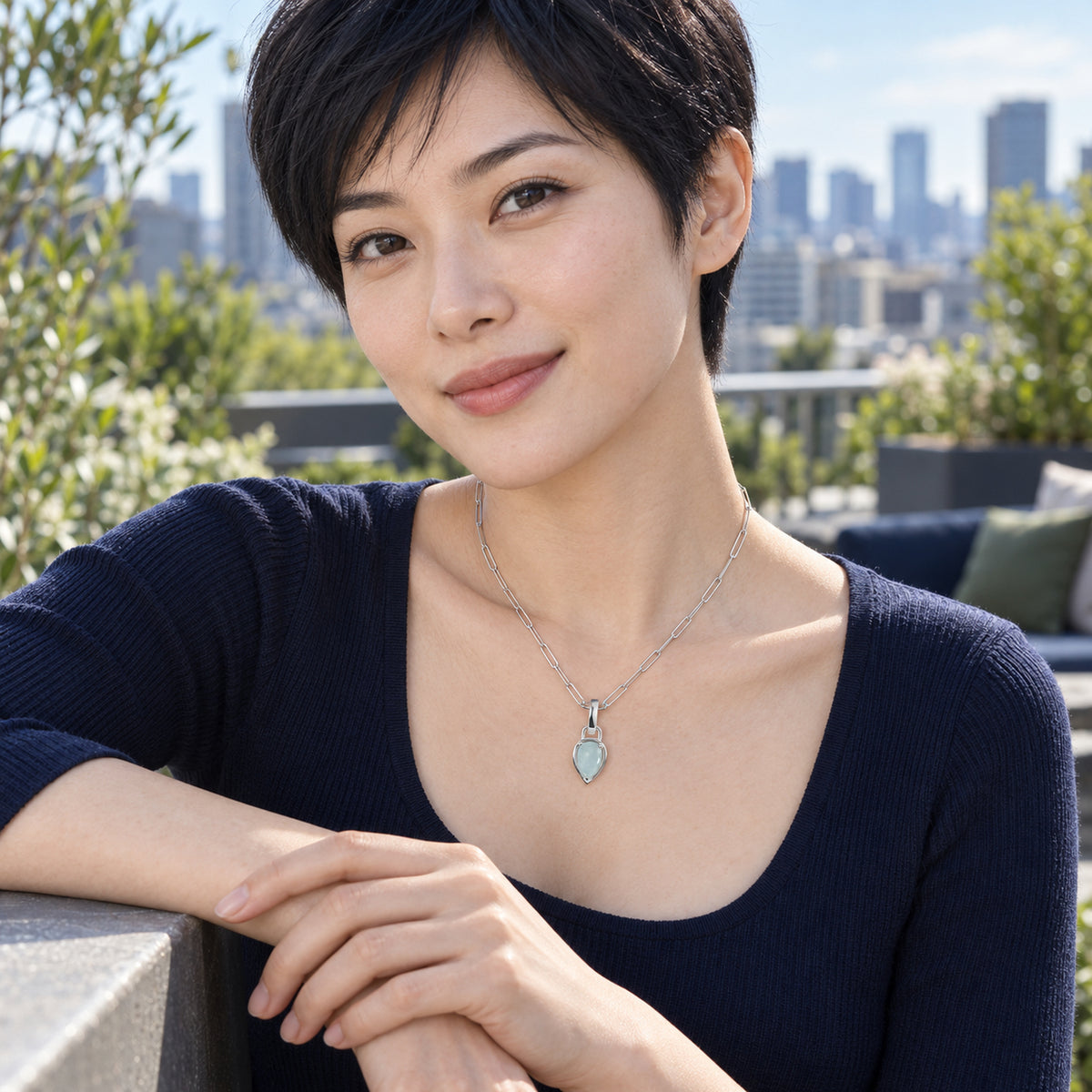 A woman with short black hair wears the silver-colored aquamarine necklace with a navy top on a terrace overlooking city buildings.