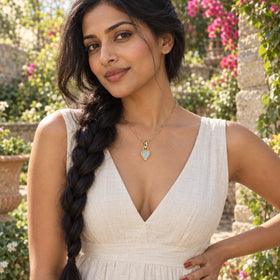 A woman with a long dark braid wears the gold-colored aquamarine pendant necklace with a white linen dress in a flowering garden.