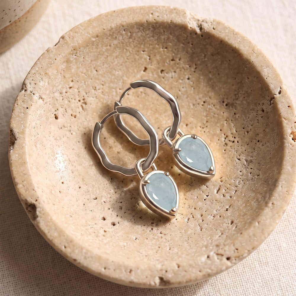 Silver-tone aquamarine hoop earrings lie diagonally together in a textured beige stone dish.