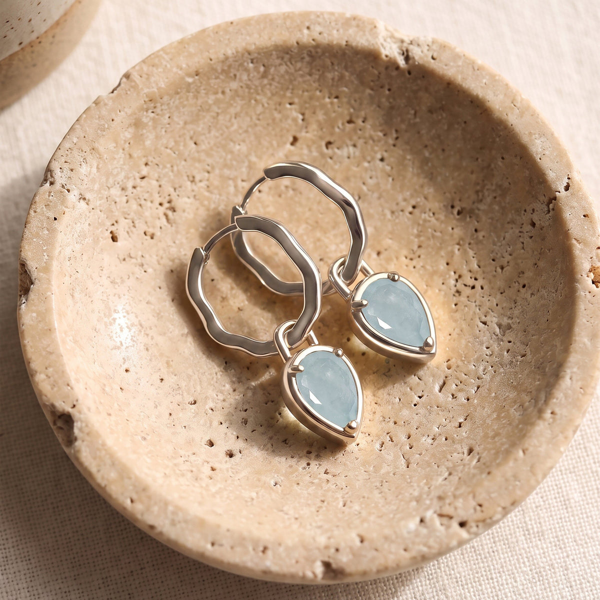 Silver-tone aquamarine hoop earrings lie diagonally together in a textured beige stone dish.