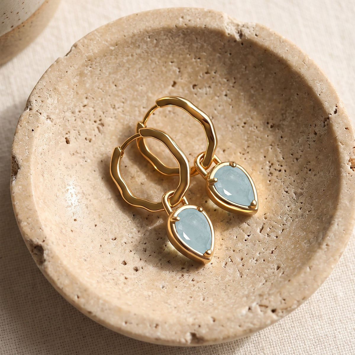 Yellow gold-tone aquamarine hoop earrings are arranged on a shallow stone dish over cream fabric.