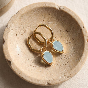 Yellow gold-tone aquamarine hoop earrings are arranged on a shallow stone dish over cream fabric.