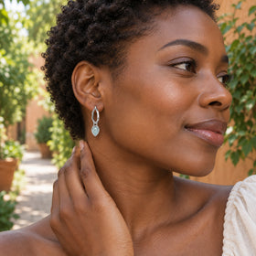 A woman with short curls wears a silver-tone aquamarine drop hoop beside a leafy garden path.