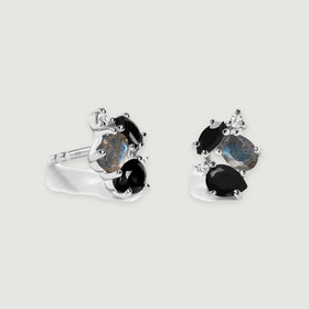 A pair of sterling silver stud earrings features black faceted stones, blue gray oval stones and small clear accents on a light gray background.