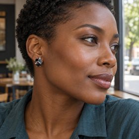 A woman with short natural curls wears a sterling silver stud earring with black and blue gray stones beside a cafe window.