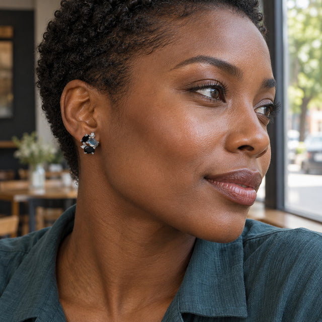 A woman with short natural curls wears a sterling silver stud earring with black and blue gray stones beside a cafe window.