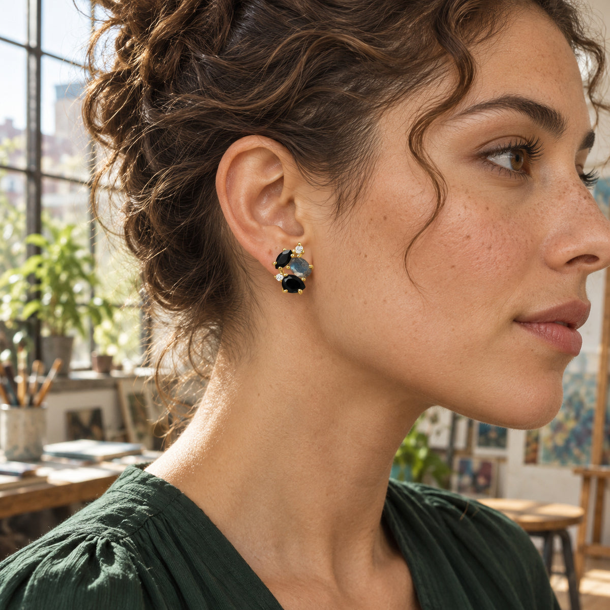 A woman with chestnut curls wears a yellow gold vermeil stud earring with black and blue gray stones in a sunlit studio.