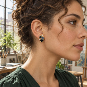 A woman with chestnut curls wears a yellow gold vermeil stud earring with black and blue gray stones in a sunlit studio.