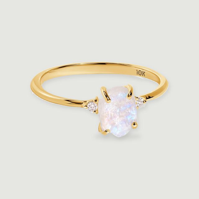 A yellow gold ring with a raw pale moonstone and two round diamond accents rests on a light gray surface.
