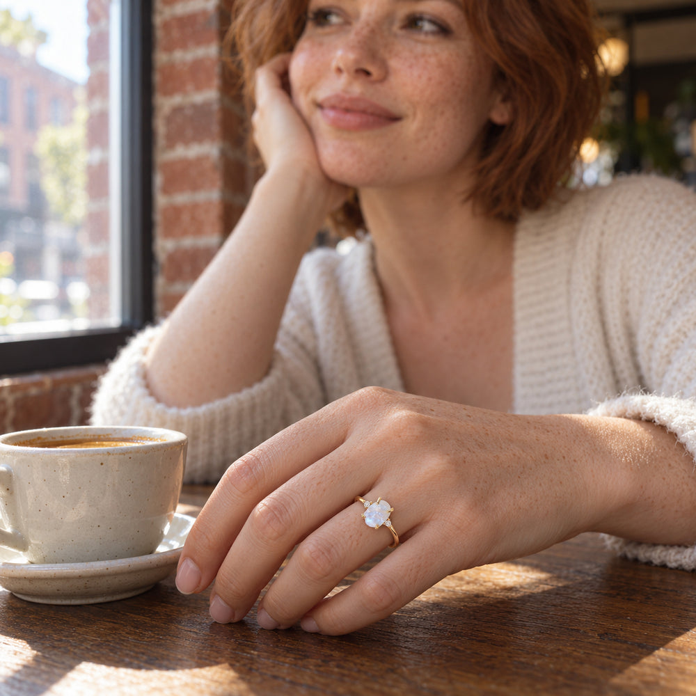 A yellow gold moonstone ring is worn on a freckled hand beside a ceramic coffee cup in a cafe.