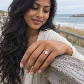 A rose gold moonstone ring is worn on a hand resting on a weathered wooden rail by the sea.