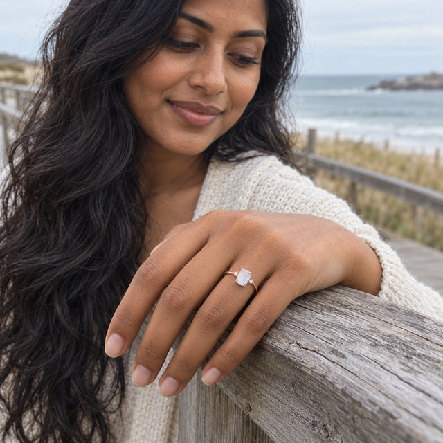 A rose gold moonstone ring is worn on a hand resting on a weathered wooden rail by the sea.
