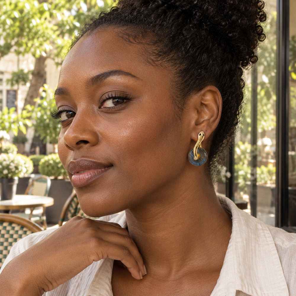 A gold-tone labradorite earring is worn beside an exposed ear in a leafy cafe courtyard.