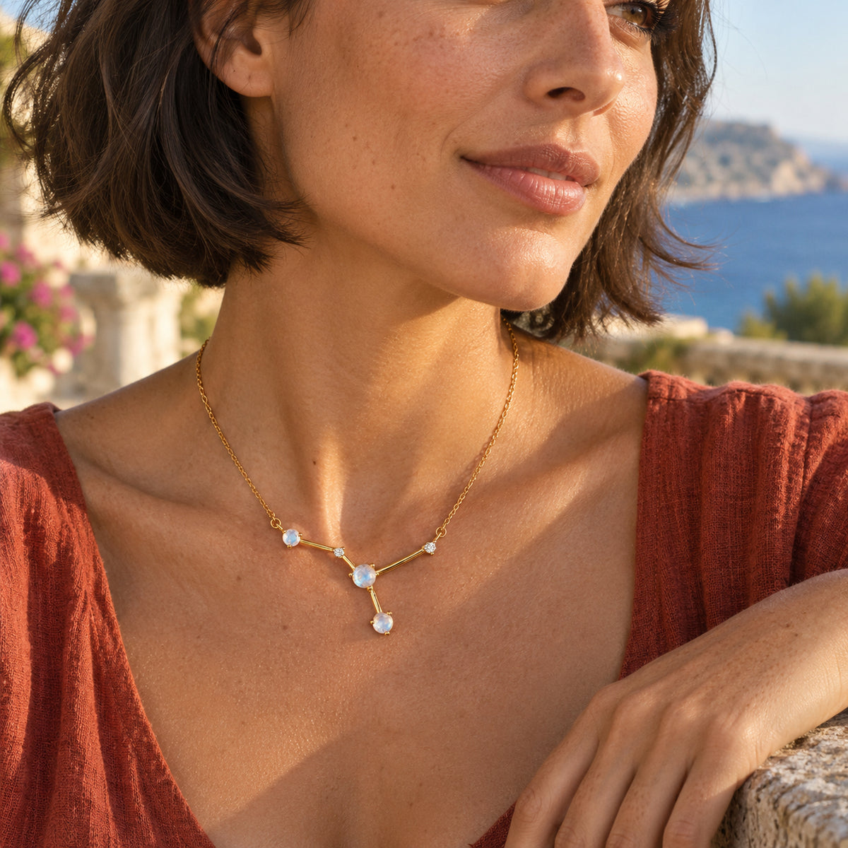 A woman with a short brunette bob wears the yellow gold necklace on a coastal terrace beside a stone railing.