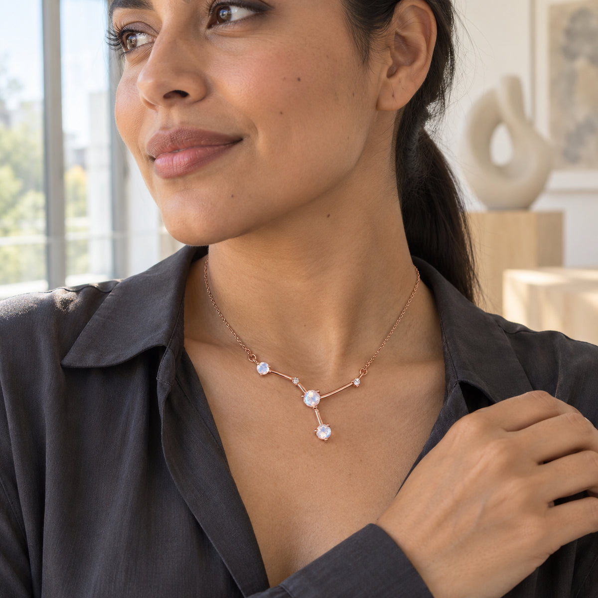 A woman wearing a charcoal blouse displays the rose gold necklace beside a bright window with pale sculptures behind her.