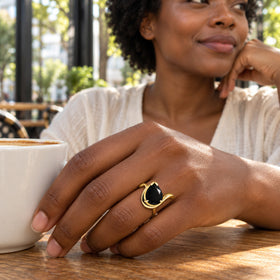 A yellow gold ring with a black pear-shaped stone is worn on a hand beside a coffee cup in a sunlit cafe.