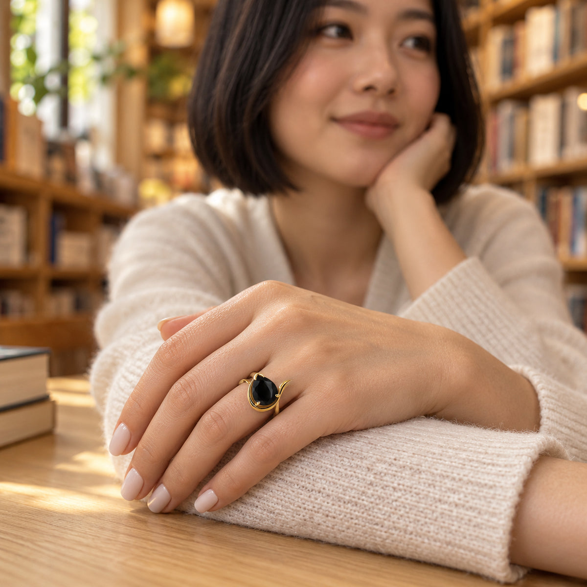 The black stone and curved yellow gold band are shown on a hand resting on a cream sleeve in a bookshop.