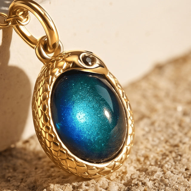 The gold-toned pendant is seen close up with a domed teal stone and engraved details beside a cream stone block.