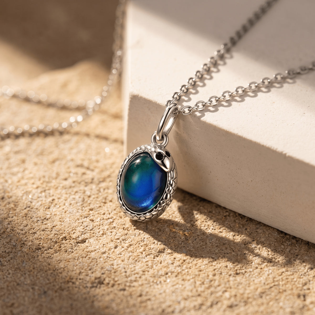 A silver-toned chain drapes across a pale stone block with its oval pendant resting on a sandy surface.
