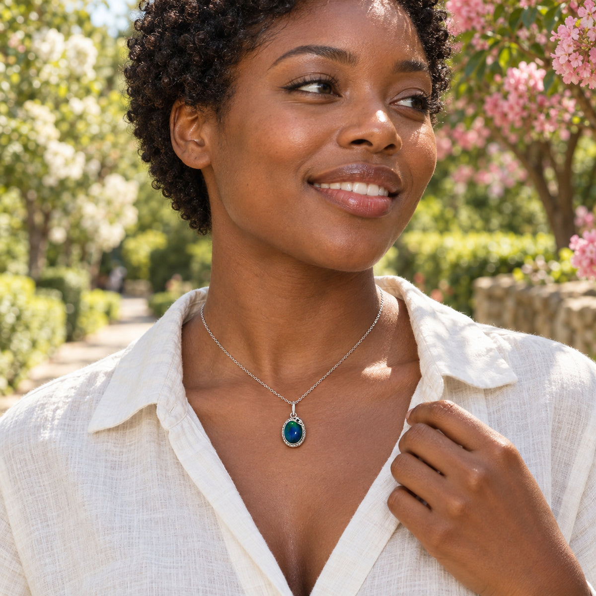 A woman in an ivory linen shirt wears a silver-toned oval blue-green pendant beside a flowering garden path.
