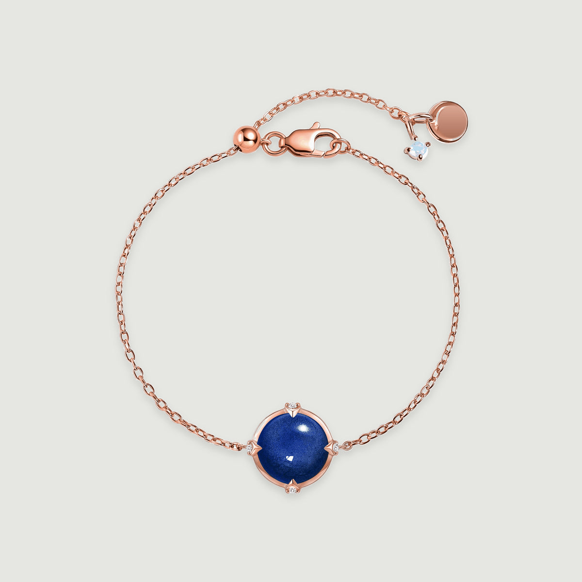 A rose gold tone bracelet with a glossy blue cabochon, delicate links, and a lobster clasp rests on pale gray.