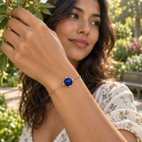 A woman holds a leafy branch in a garden while wearing a yellow gold tone bracelet with a round blue stone.