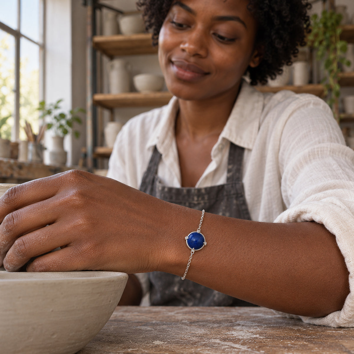 A silver tone blue stone bracelet rests on a woman's wrist beside a ceramic bowl in a pottery studio.
