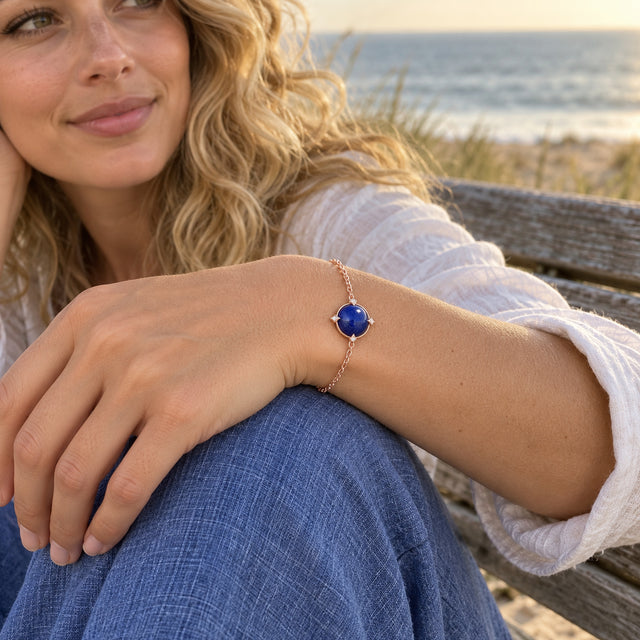 A rose gold tone bracelet with a blue cabochon is worn above a relaxed hand resting on blue fabric by the sea.