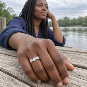 A white gold moonstone ring is worn on a hand resting on weathered wood beside a lake.