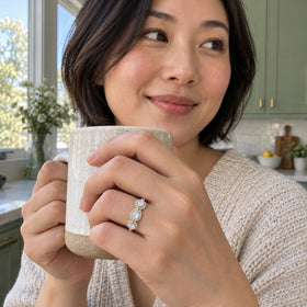 A yellow gold ring with three pale stones is shown on a hand holding a ceramic cup in a sunlit kitchen.
