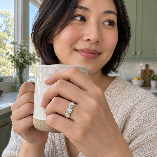 A yellow gold ring with three pale stones is shown on a hand holding a ceramic cup in a sunlit kitchen.
