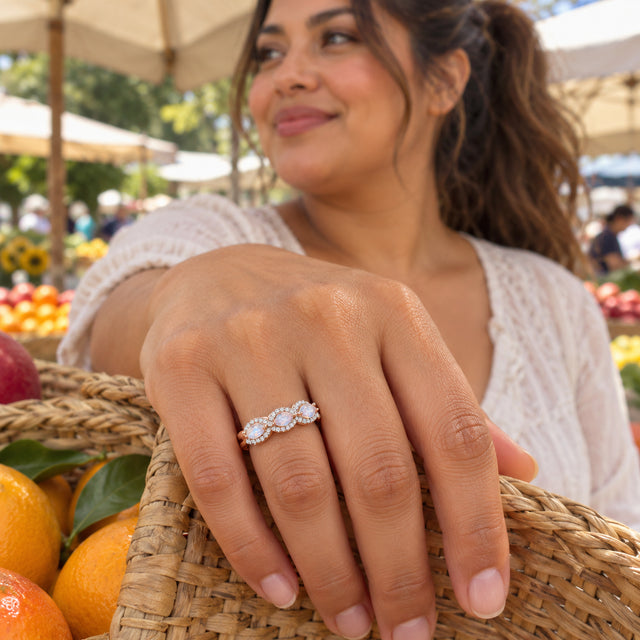 A rose gold ring with diamond framed moonstones is worn above a woven basket of fruit.