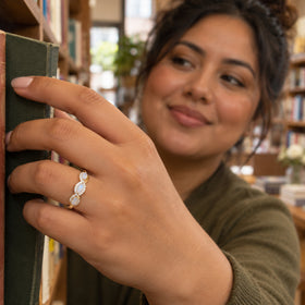 A hand wearing a yellow gold ring with three pale moonstones rests against a green book in a bookstore.