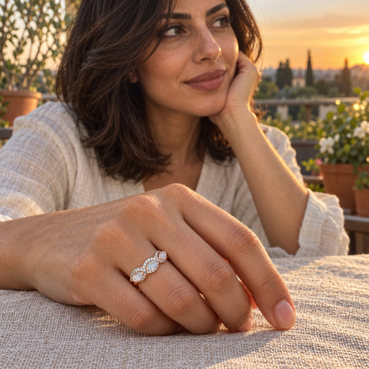 A rose gold moonstone ring is shown on a hand resting on a linen tablecloth beside a garden balcony at sunset.