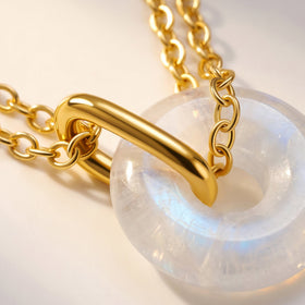 Yellow gold vermeil chain links and a curved connector cross a translucent moonstone on a cream tabletop.
