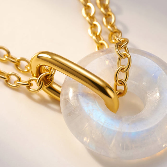Yellow gold vermeil chain links and a curved connector cross a translucent moonstone on a cream tabletop.